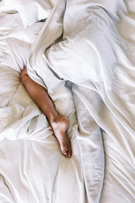 A single leg and foot extending out from under crisp white bedding. The image shows a rumpled, unmade bed with white sheets and duvet, with just one leg visible, suggesting someone is still sleeping or relaxing under the covers.