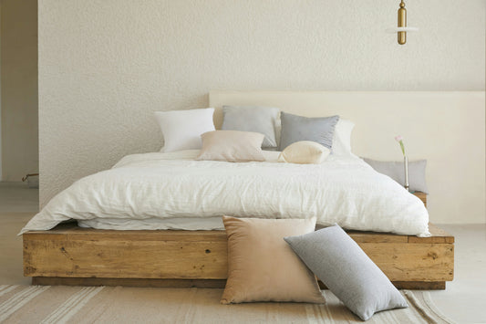 Minimalist bedroom featuring a low wooden platform bed with white linen bedding, neutral-toned throw pillows in beige and gray, and a simple pendant light against textured white walls