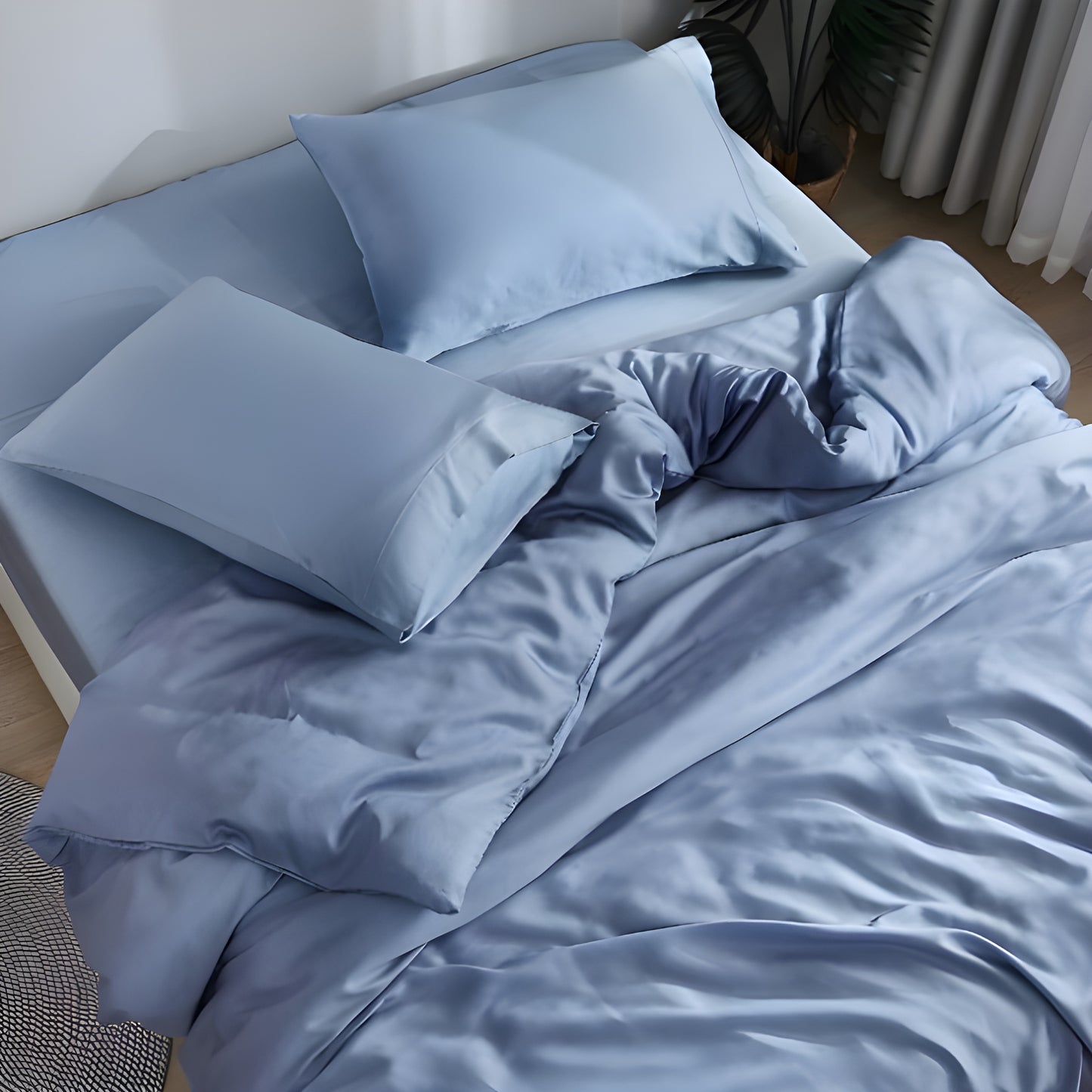 Luxurious light blue bamboo bedding set displayed on a modern bed frame in a bright, minimalist bedroom. The silky bedding features a smooth, lustrous finish that captures light beautifully, creating an elegant sheen across the slightly rumpled duvet cover and pillowcases. Two plush pillows rest against the headboard, complemented by the flowing sheets.