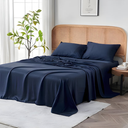 Luxurious navy blue bedding set elegantly styled on a bed with a natural rattan headboard in a bright, airy bedroom. The premium sheets showcase their smooth, flowing drape with subtle folds that highlight the fabric's exceptional quality. Two matching navy pillowcases rest against the textured headboard, creating a striking contrast.